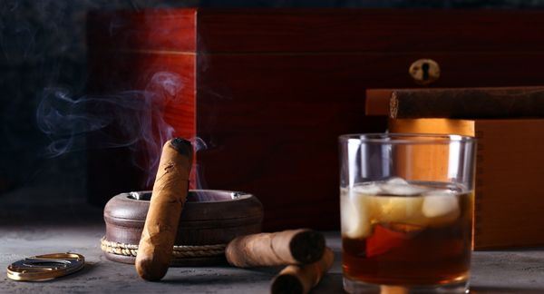 Cigar burning on ashtray next to boxes and bourbon glass