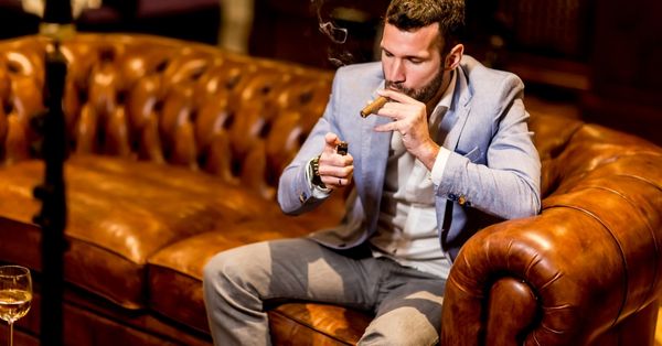 Man in blue blazer lighting a premium cigar on a brown leather sofa